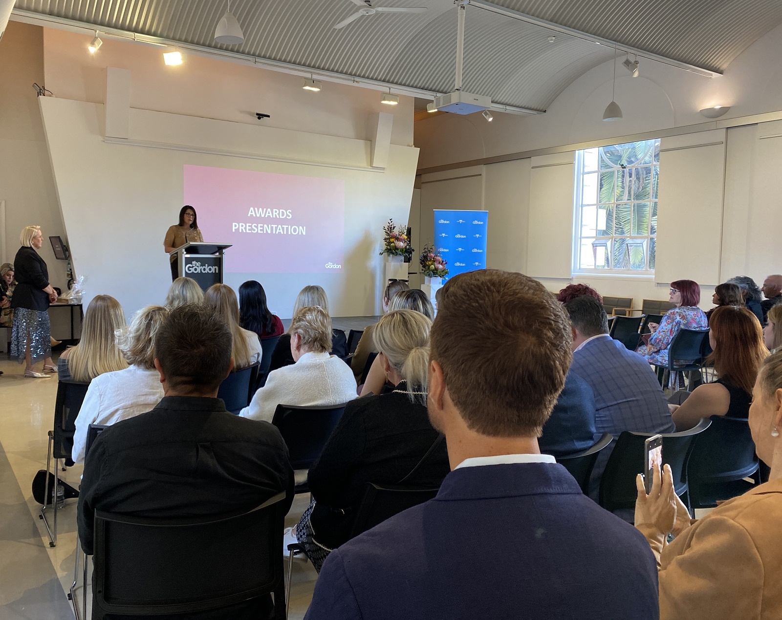 Image of a group of people attending, seated, watching a person at a podium presenting an award.