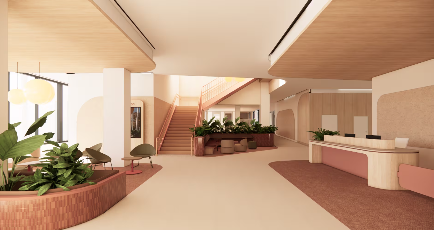 Centre of Excellence Entrance. A light-filled building foyer with a central staircase, surrounded by indoor plants and seating. A reception desk sits to the right, with open walkways and soft pink and neutral tones throughout the space.