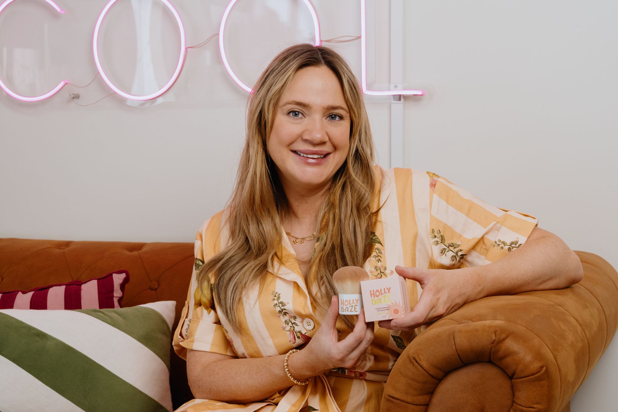 Image of Kayla Houlihan sitting on a couch holding some of their Holly Daze skin care products.