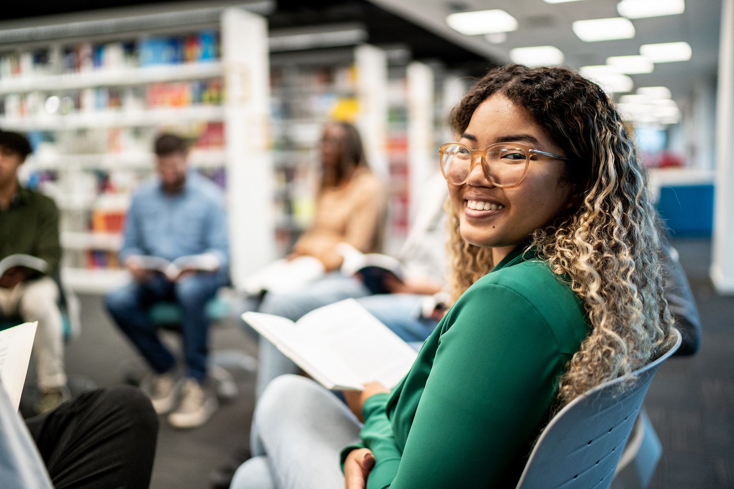Image of a student in the library reading a boook. They are reclining and smiling.